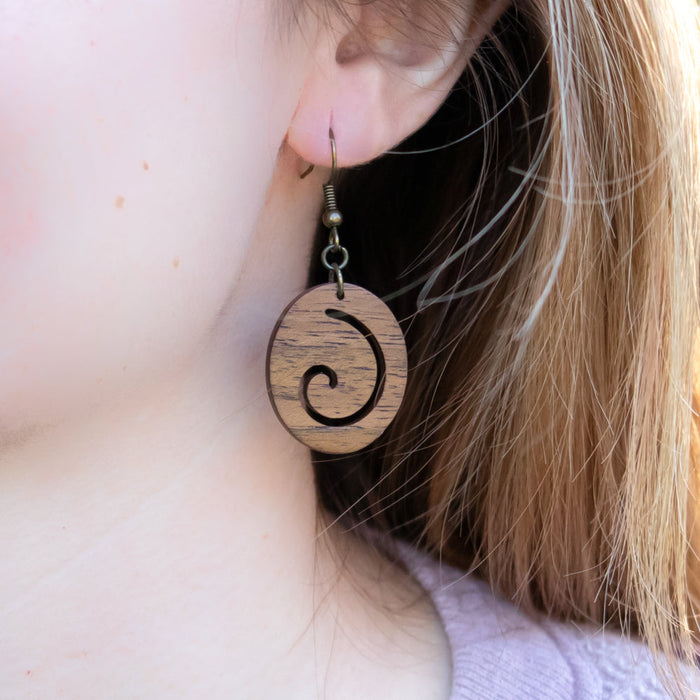 This pair of smoothed wooden drop earrings features an abstract spiral within an ovoid shape. Available in a red oak, walnut, or cherry.  Each piece is handmade and unique. Placement and colors may vary and are not guaranteed but will resemble the piece shown.      1" long x 0.75" wide     Ear hook is bronze and nickle free.